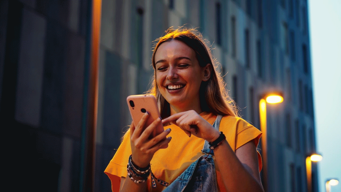 GiF - Junge Frau steht zwischen Hochhäusern und schaut lächelnd auf ihr Smartphone