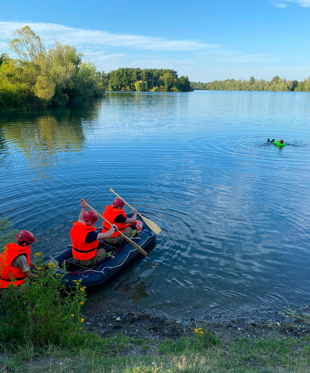 Ein Schlauchboot mit drei Feuerwehrleuten, die auf einem See zu einem Kollegen paddeln, welcher einen Überlebensanzug trägt