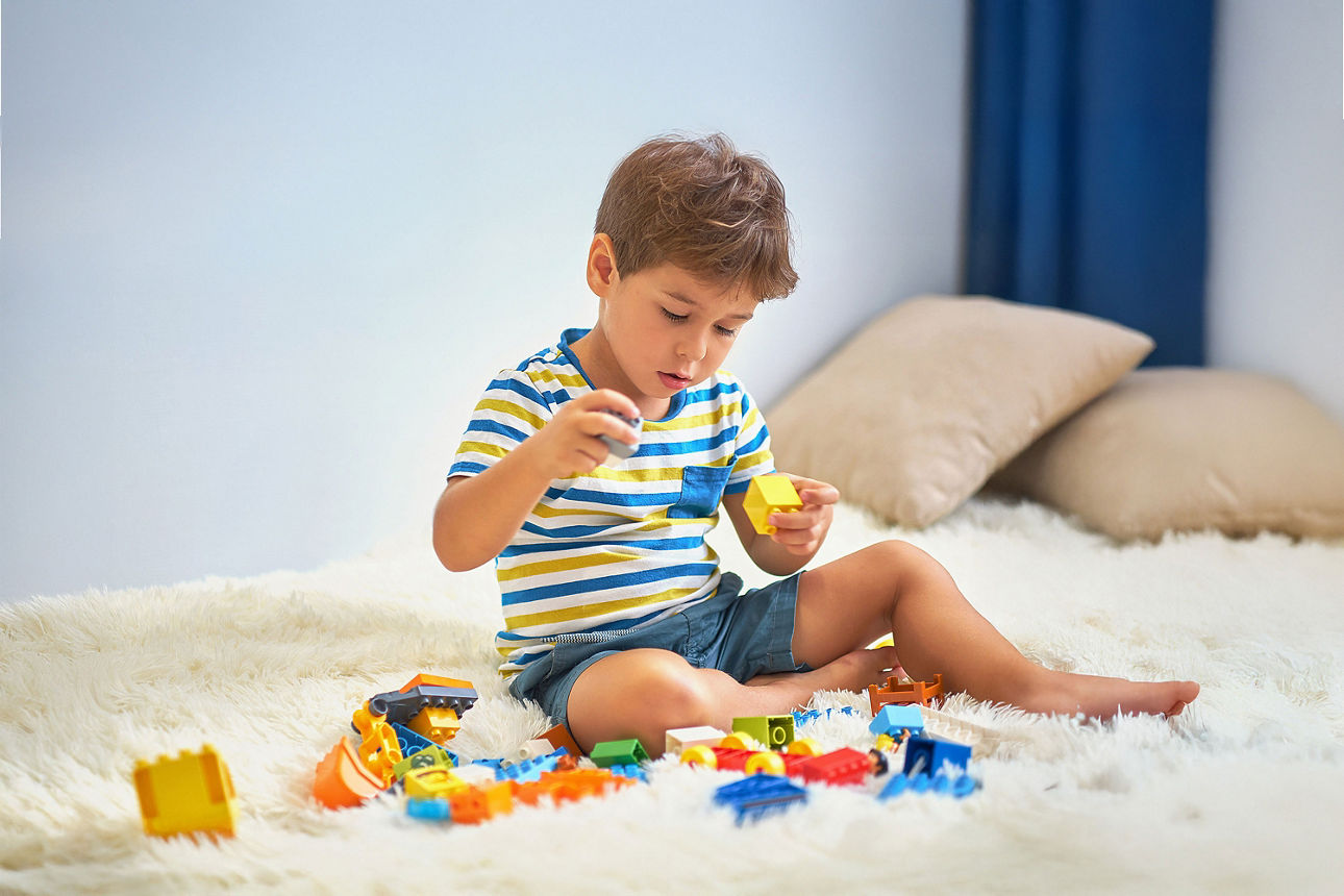 Ein kleiner Junge im gestreiften T-Shirt sitzt auf einem flauschigen weißen Teppich und spielt konzentriert mit bunten Bauste