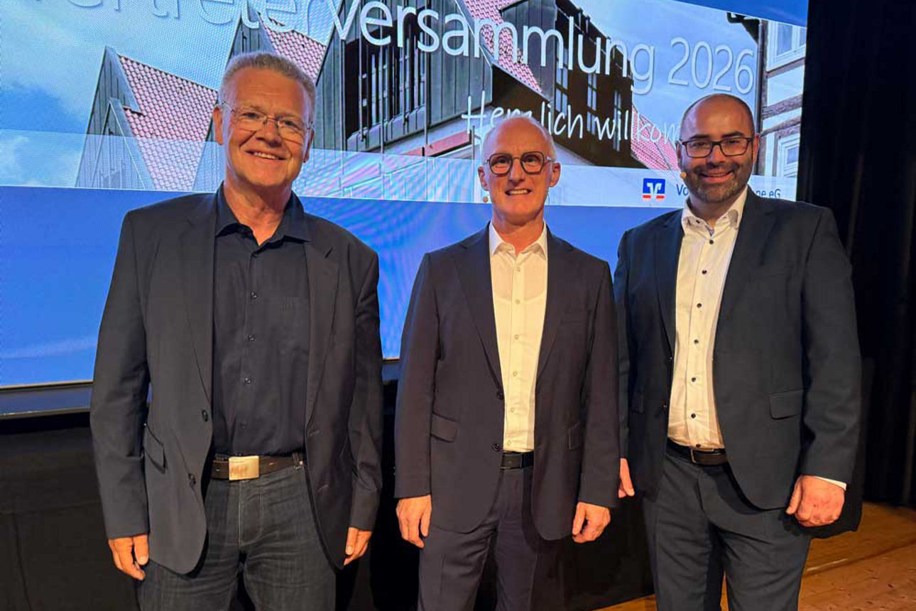 Drei Männer in eleganten Anzügen posieren lächelnd auf einer Bühne vor einer großen Leinwand mit der Aufschrift „Vertreterver