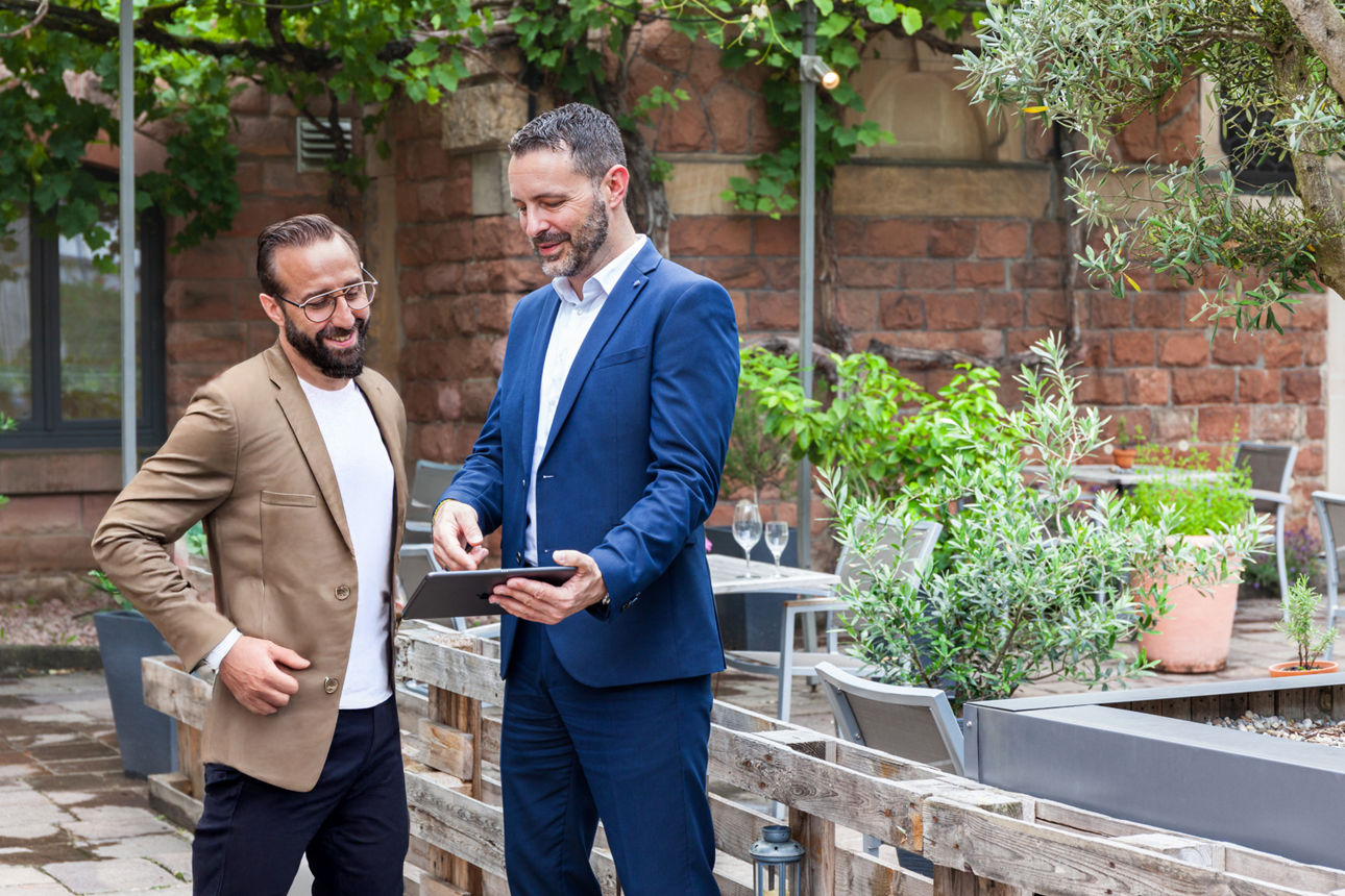 Zwei Personen im Gespräch im Freien, eine im Anzug mit Tablet, die andere in Freizeitkleidung