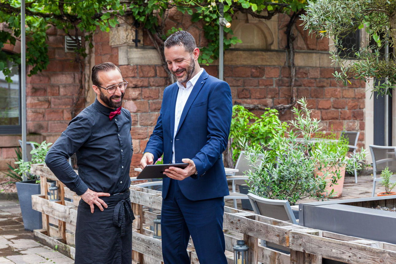 Ein Mann im Anzug zeigt einem Kellner mit Fliege und Schürze ein Tablet. Sie stehen in einem begrünten Außenbereich