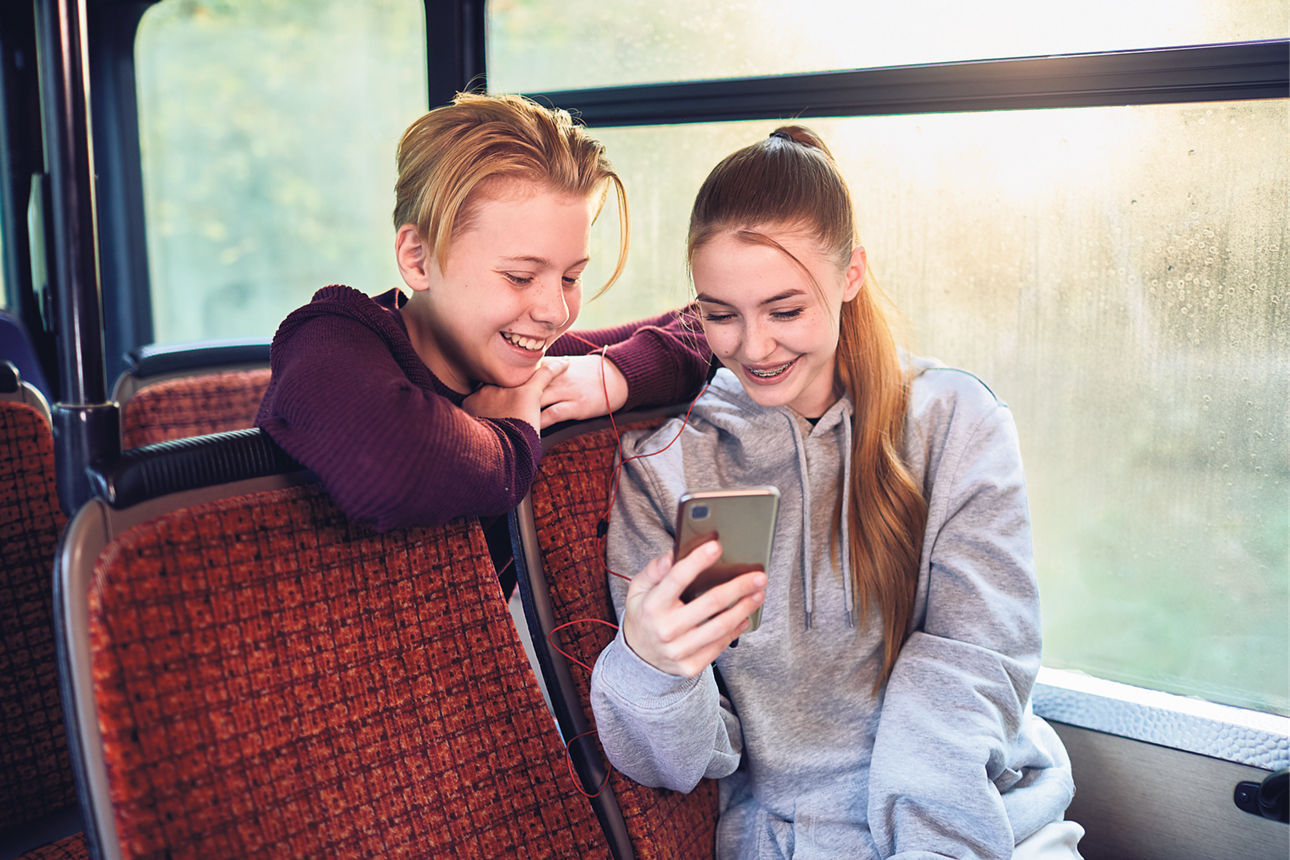 Zwei Kinder sitzen im Bus und schauen gemeinsam auf ein Smartphone. 