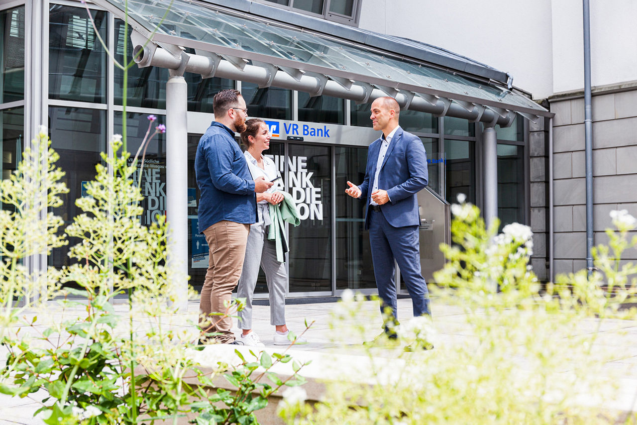 Drei Personen im Gespräch vor einer VR-Bank-Filiale, moderne Glasfassade im Hintergrund, Pflanzen im Vordergrund.