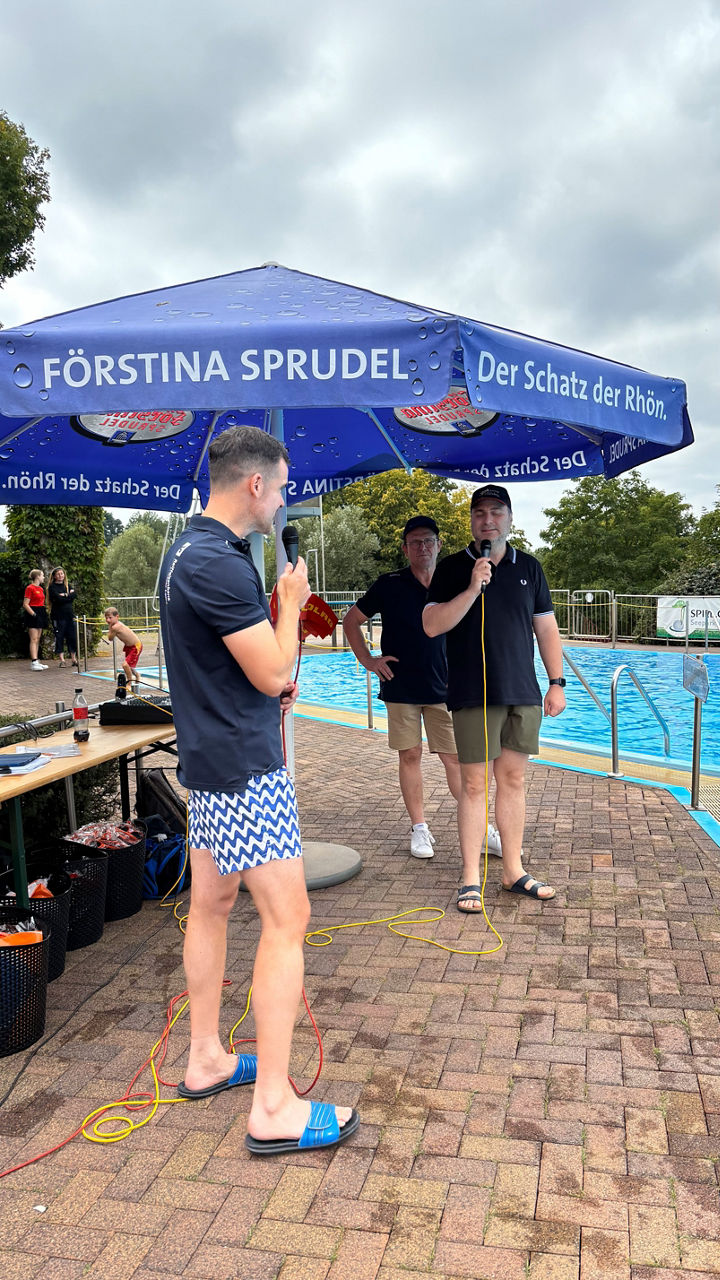 Veranstaltungsmoderator spricht in ein Mikrofon am Rand eines Swimmingpools.
