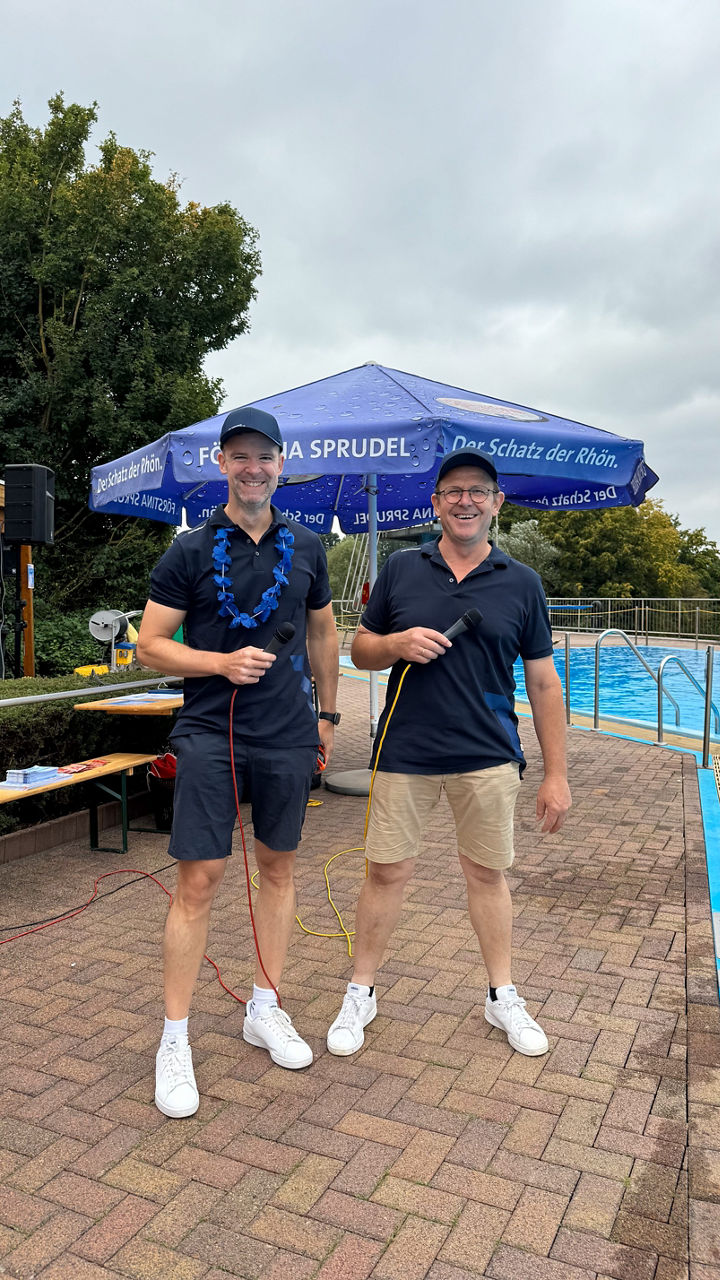 Zwei Männer mit Mikrofonen in der Hand stehen bereit an einem Pool unter einem blauen Sonnenschirm.