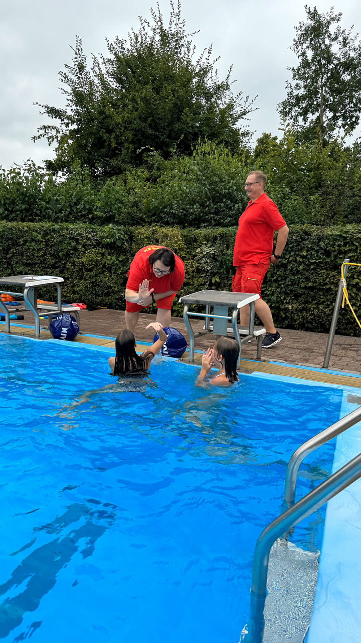 Rettungsschwimmer leiten Schwimmer in einem strahlend blauen Pool unter bewölktem Himmel, mit grünen Bäumen im Hintergrund.