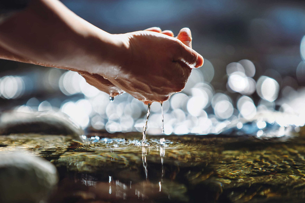 Ausschnitt eines Fotos von zwei Händen die klares Wasser aus einer Quelle schöpft. 