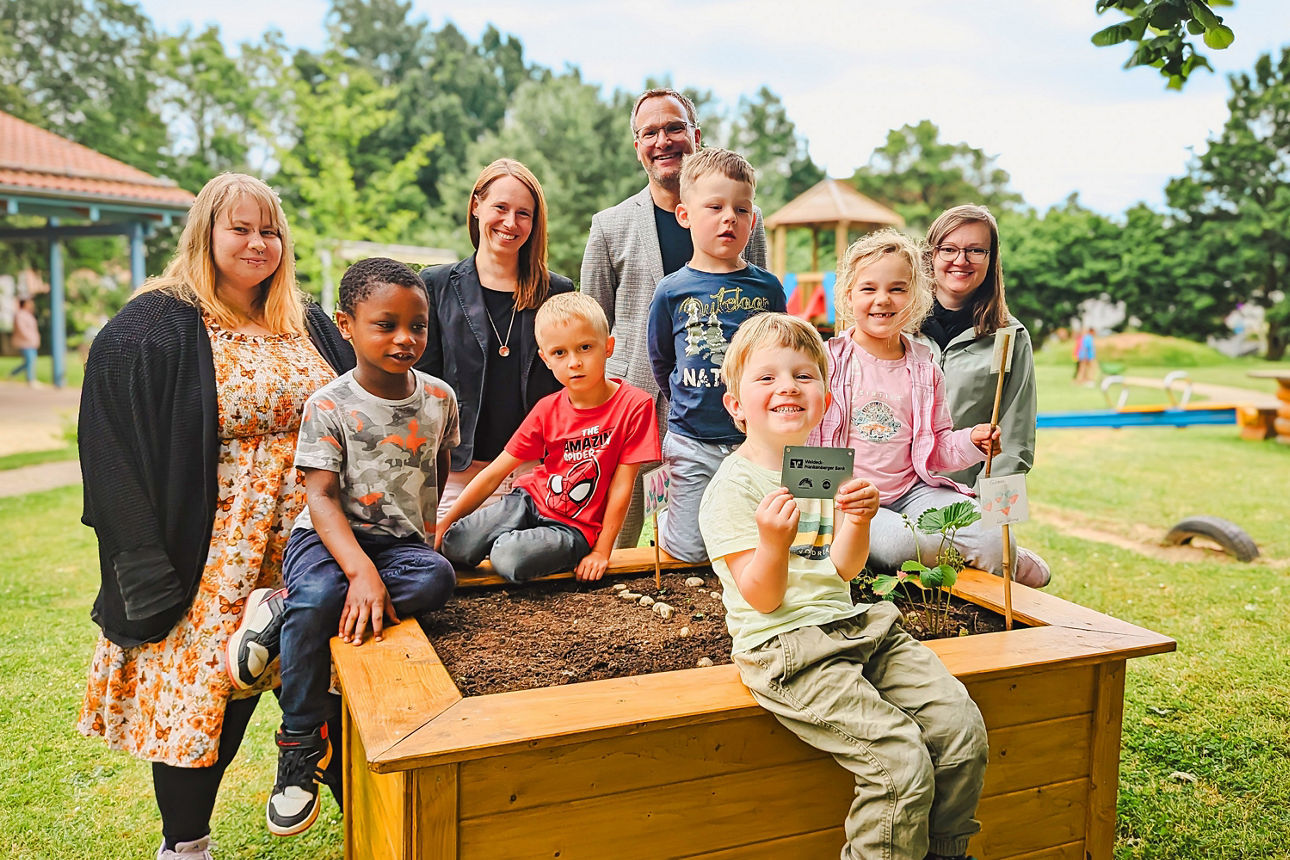 Eine Familie steht zusammen und lächelt vor einem Blumenbeet, das mit Pflanzen und Blumen gefüllt ist.