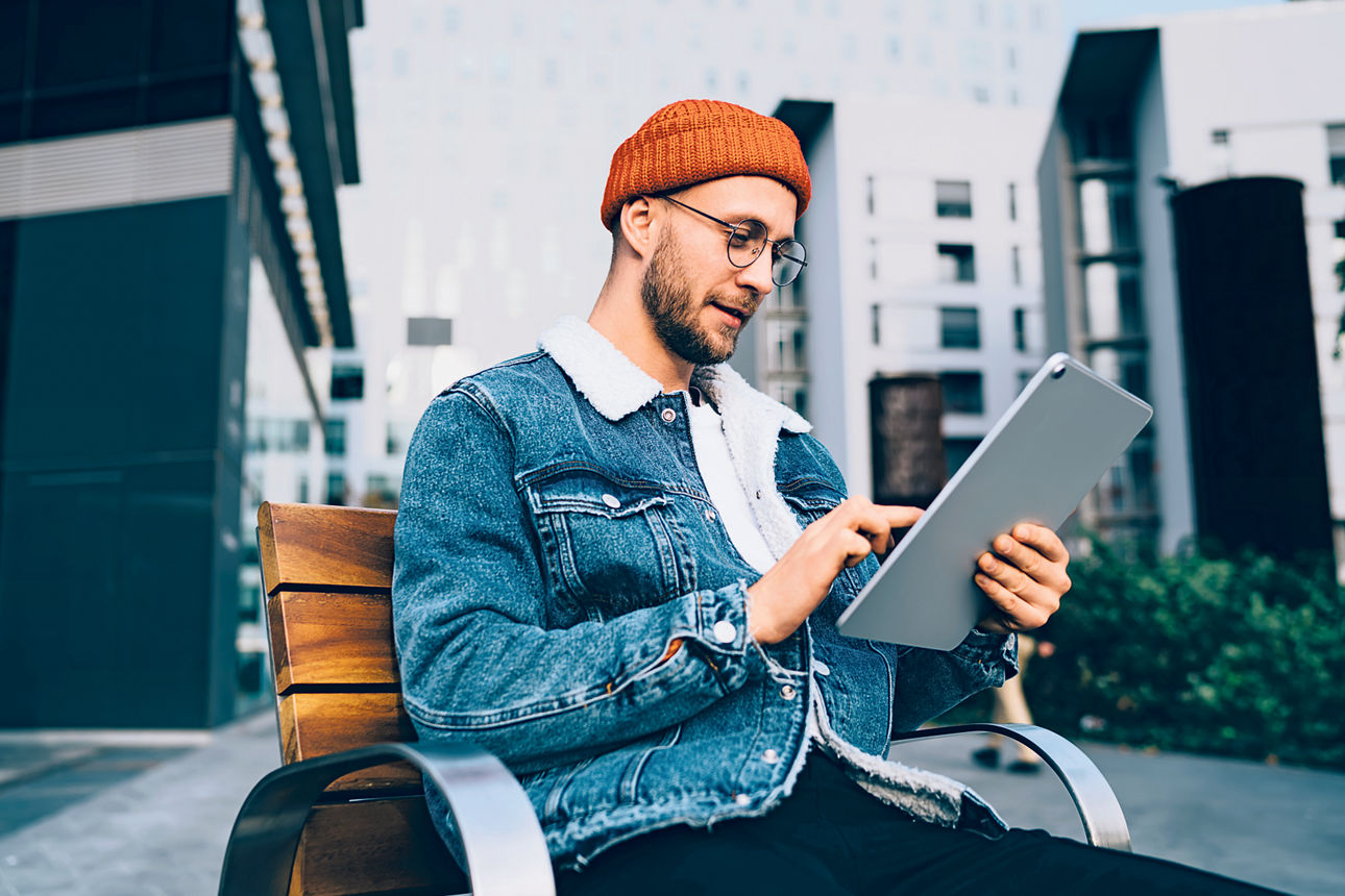 Ein Mann mit Brille, orangefarbenem Hut und Jeansjacke sitzt auf einer Bank vor einigen Gebäuden