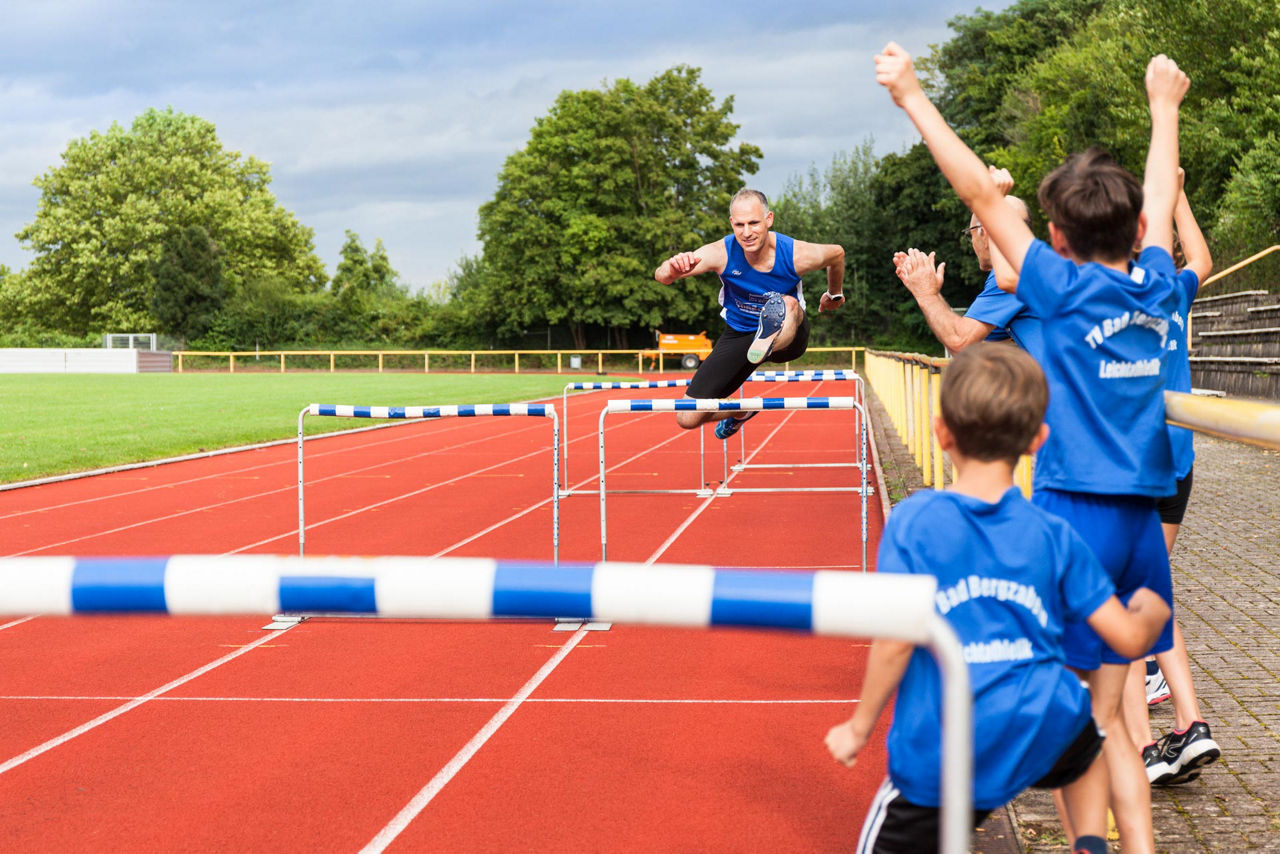 Athlet springt über Hürden auf Laufbahn, Kinder am Rand jubeln.