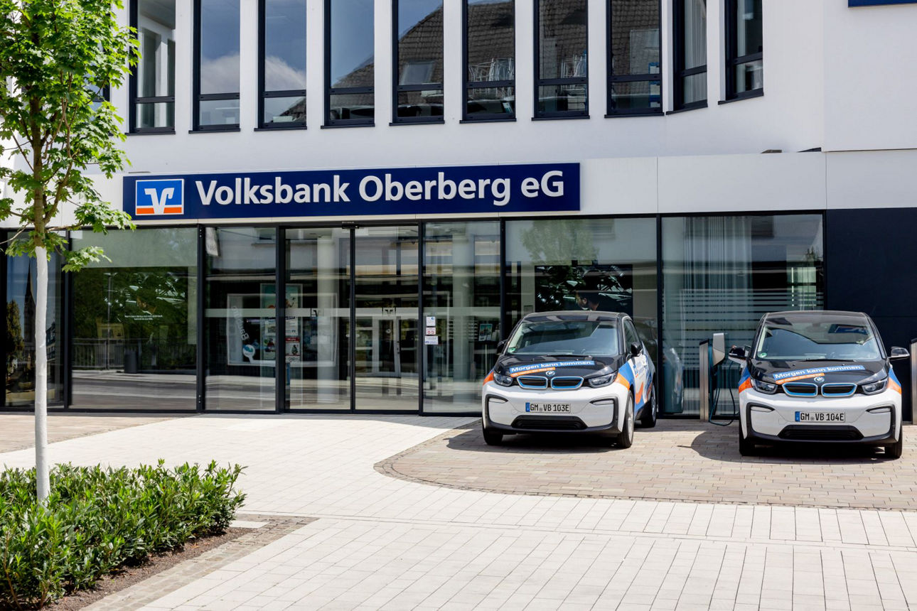 Eingang der Volksbank Oberberg eG mit Glasfront, ebenerdigem Zugang und zwei geparkten Elektroautos.
