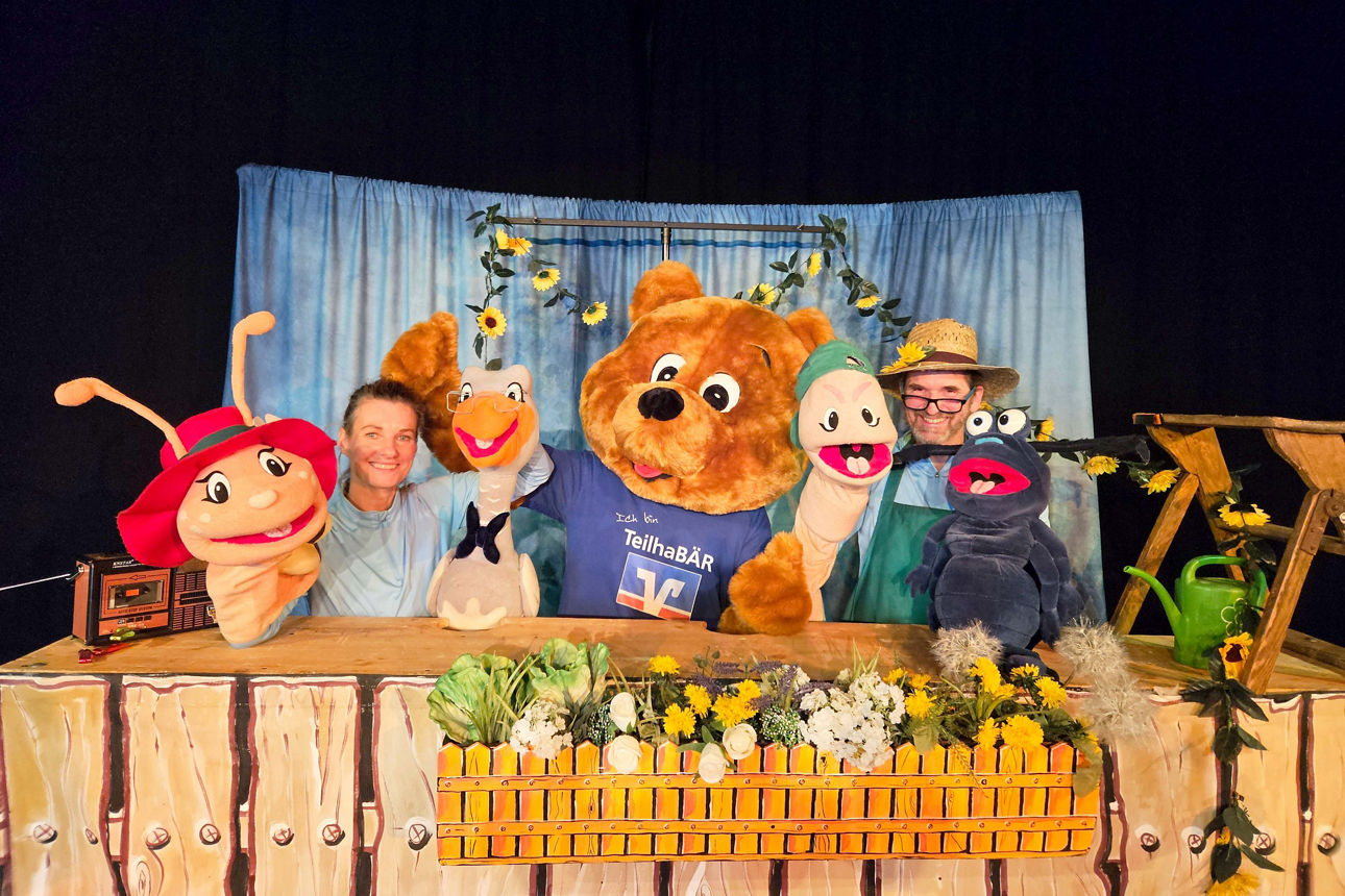 Maskottchen der VR-Bank Mittelsachsen steht zwischen einem Mann und einer Frau mit Handpuppen