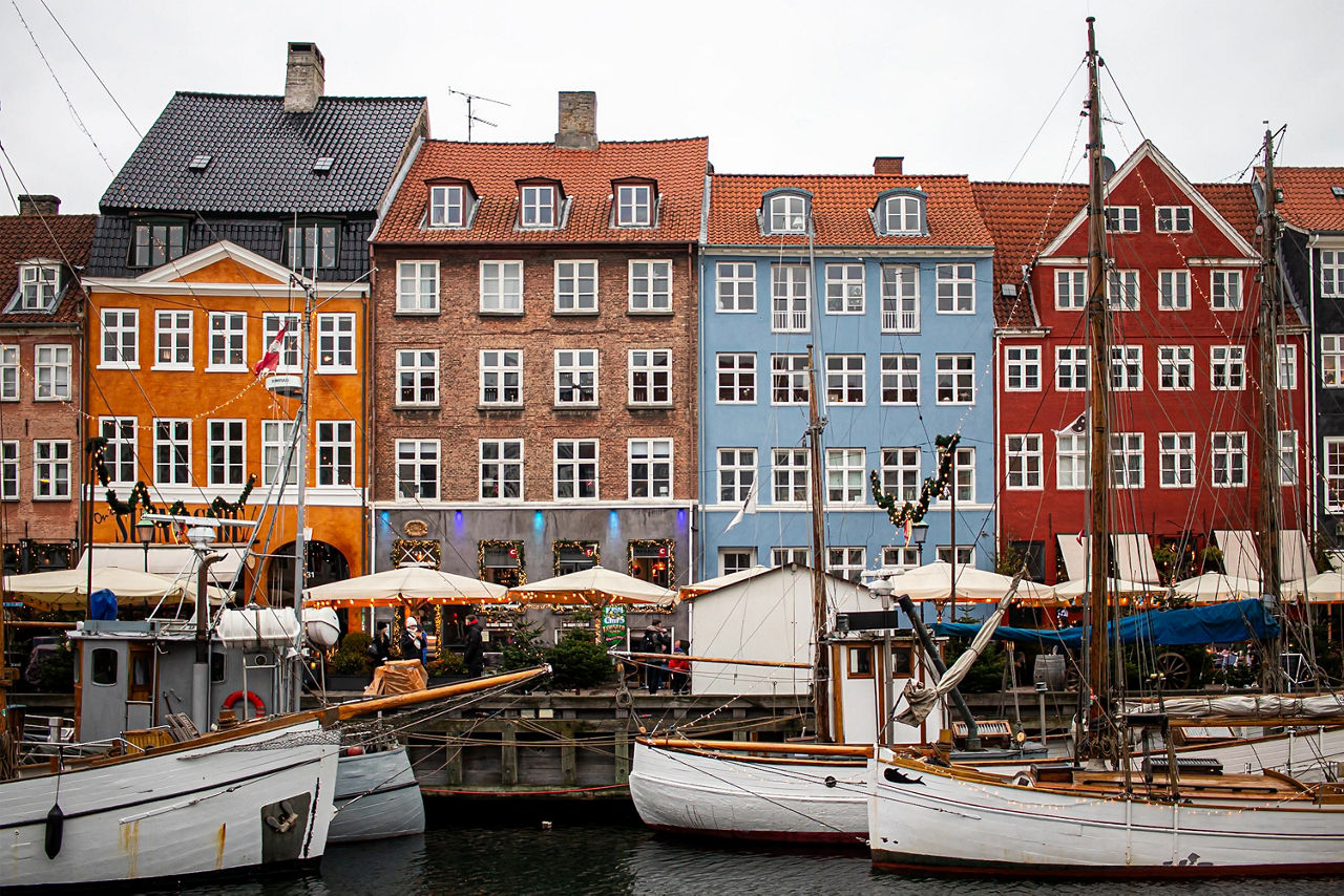 Bunte, mehrstöckige Häuserreihe am Wasser mit mehreren Segelbooten im Vordergrund und Weihnachtsbeleuchtung.