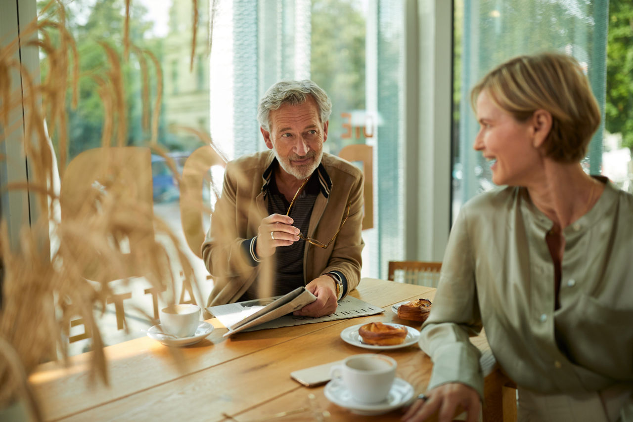 Ein älteres Pärchen sitzt in einem Café bei Kaffee und süßen Teilchen, der Mann hält eine Brille und eine Zeitung in der Hand