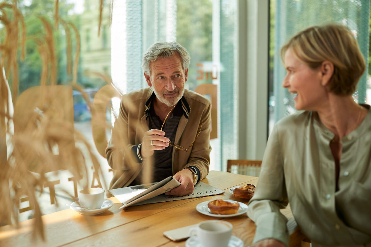 Ein älteres Pärchen sitzt in einem Café bei Kaffee und süßen Teilchen, der Mann hält eine Brille und eine Zeitung in der Hand