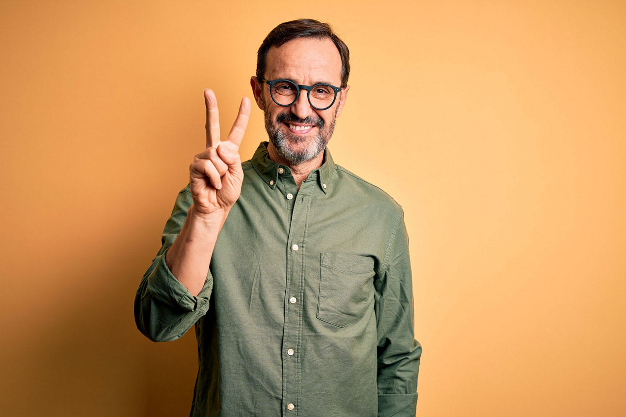 Mann mit Brille und grünem Hemd streckt zwei Finger in die Luft und lächelt in die Kamera