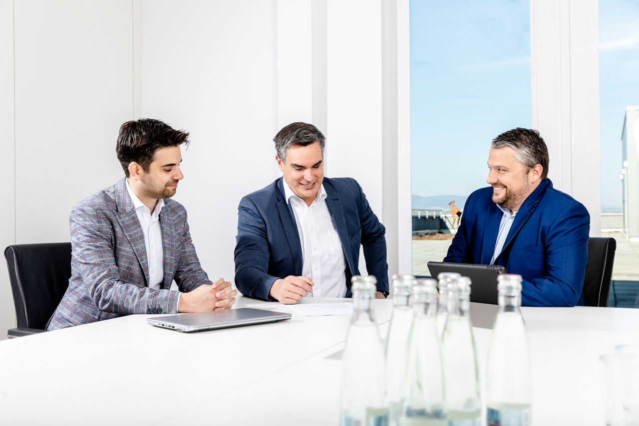 Drei Personen im Gespräch an einem Tisch mit Laptop, vermutlich in einer Beratungssituation.