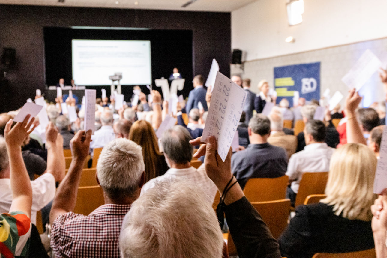 Menschen heben Stimmzettel bei einer Versammlung, im Hintergrund läuft eine Präsentation