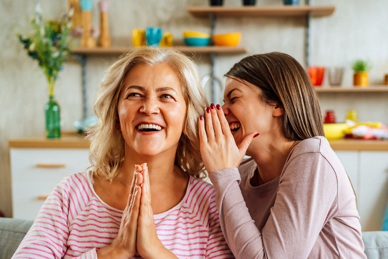 Zwei Frauen sitzten auf einem Sofa. Eine flüstert der anderen fröhlich etwas ins Ohr. 