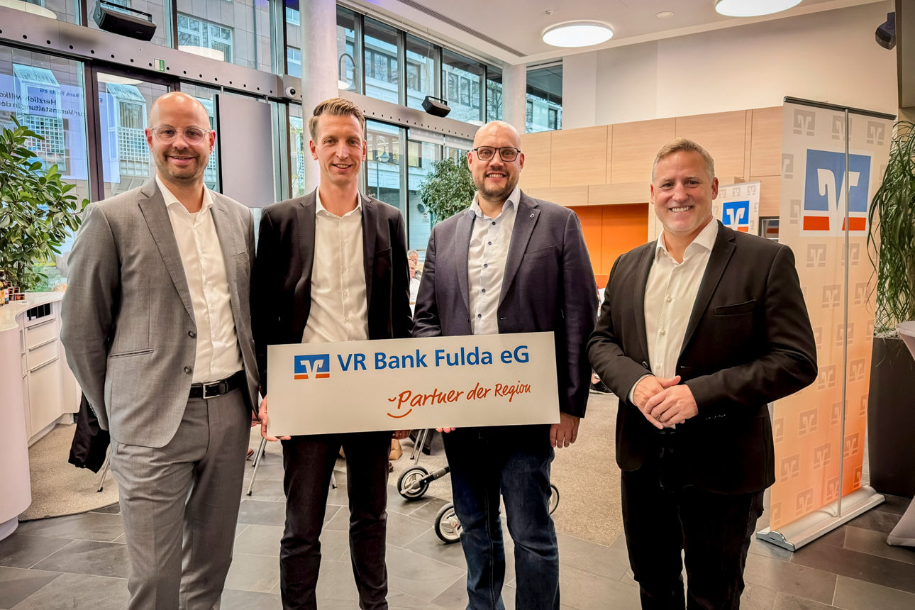 Zu sehen sind vier im Anzug gekleidete Männer. Zwei davon halten gemeinsam einen Banner, das das VR Bank Fulda Logo zeigt.