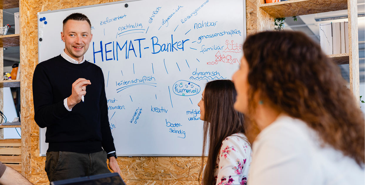Person spricht vor Whiteboard mit ‚HEIMAT-Banker‘-Titel, umgeben von aufmerksamem Publikum