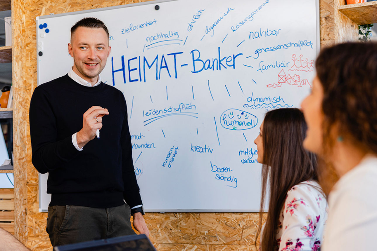 Person spricht vor Whiteboard mit ‚HEIMAT-Banker‘-Titel, umgeben von aufmerksamem Publikum