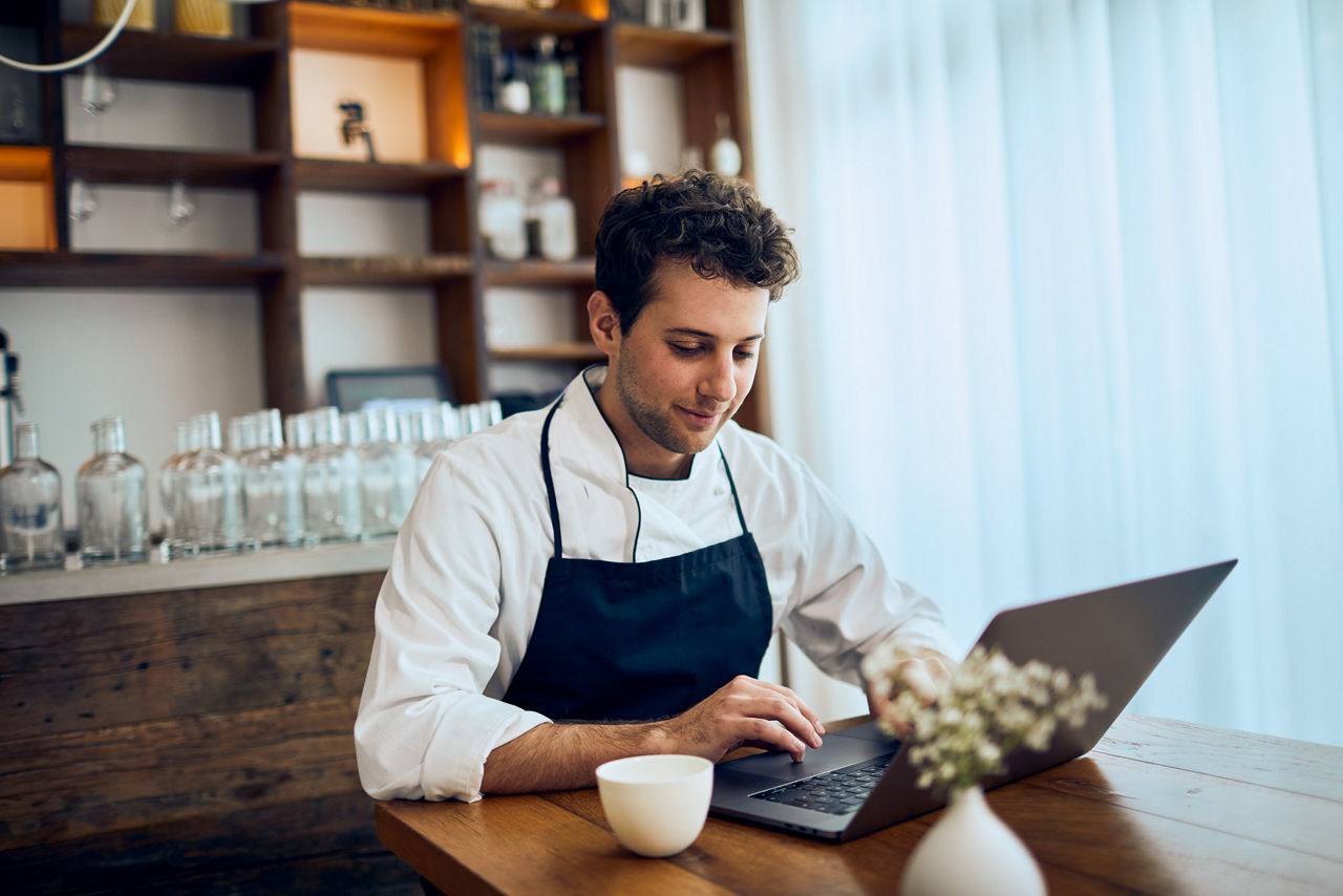 Koch sitzt an einem Tisch und checkt das Onlinebanking mit seinem Laptop