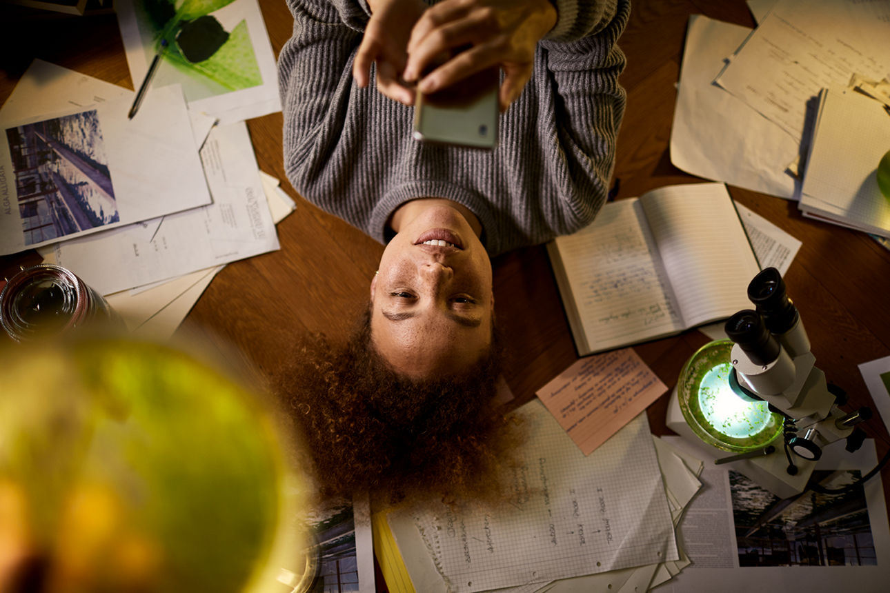 Eine Person liegt auf dem Boden, schaut auf ein Smartphone und ist von Unterlagen sowie einem Mikroskop umgeben.