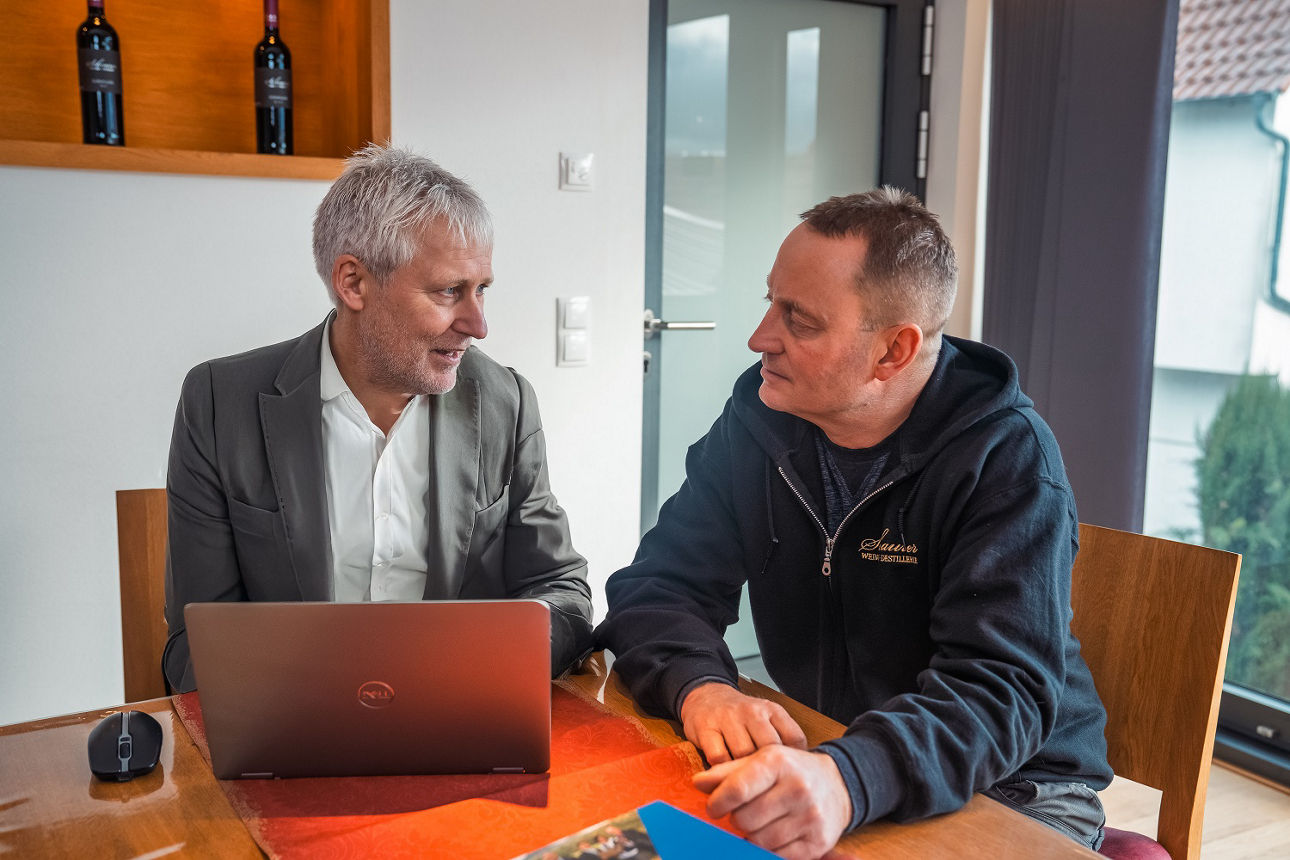Zwei Personen sitzen an einem Tisch. Vor ihnen liegt ein Laptop und einige Unterlagen.