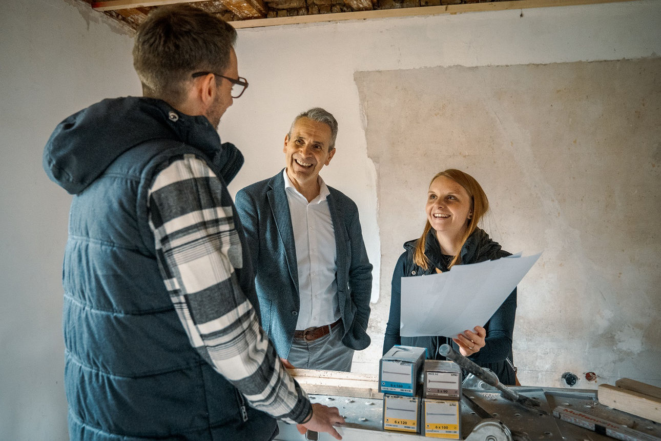 Drei Personen im Rohbau im Gespräch über Baufinanzierung, eine Person hält Unterlagen in der Hand