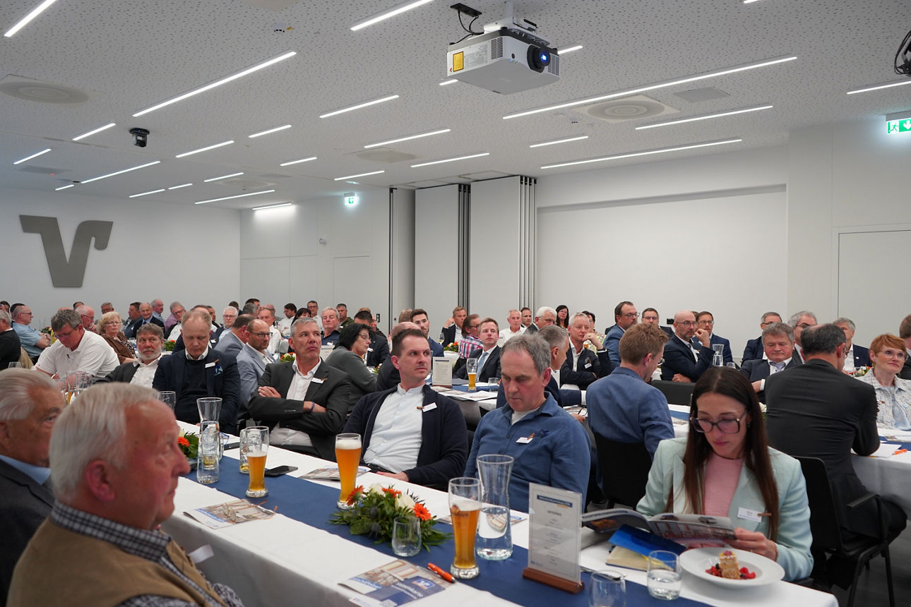 Menschen sitzen in einem Saal an langen Tischen mit Getränken vor sich