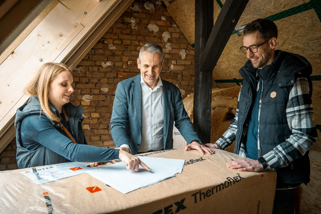Drei Personen besprechen Baupläne auf einem Dachboden mit Holzbalken und Ziegelwänden.
