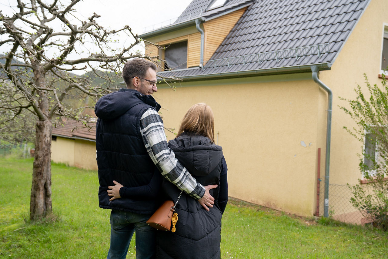 Ein Paar steht im Garten und betrachtet ein gelbes Haus mit grauem Dach und Dachfenstern.