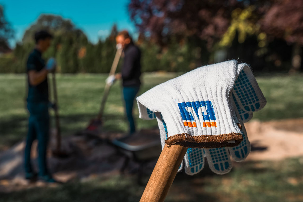 Arbeitshandschuhe mit Volksbank-Logo auf Spaten; im Hintergrund Menschen bei Garten- oder Bauarbeiten im Park.