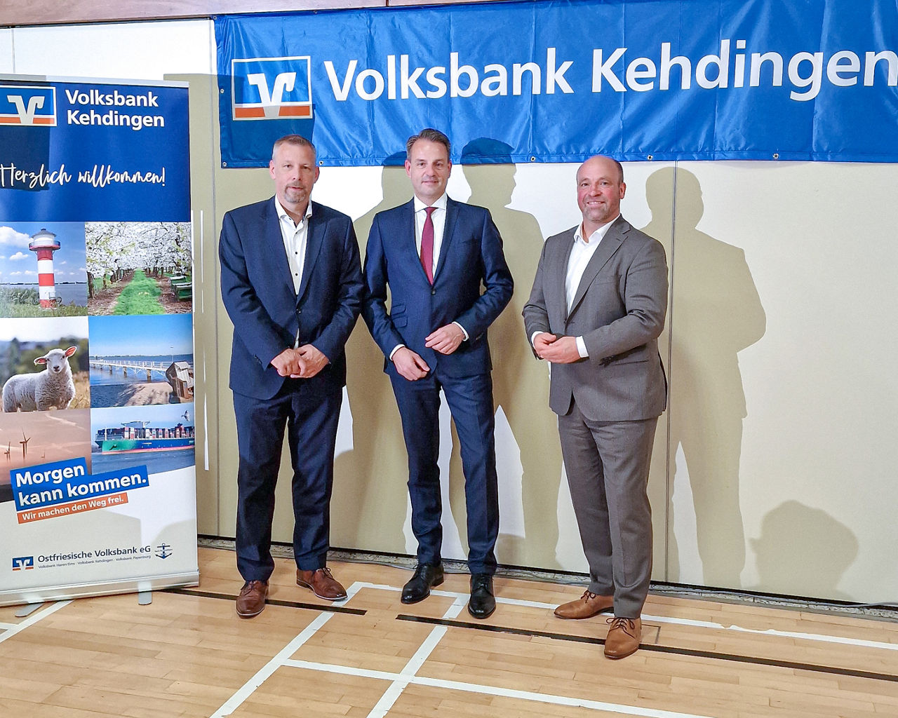 Drei Personen vor Banner der Volksbank Kehdingen, links Display mit ‚Morgen kann kommen‘-Motiv.