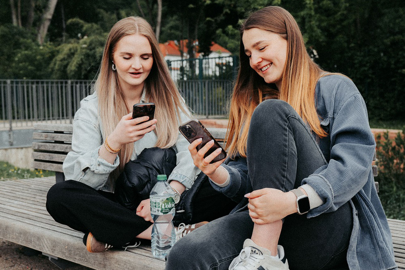 Zwei Personen sitzen auf einer Bank im Freien und schauen auf ihre Smartphones.