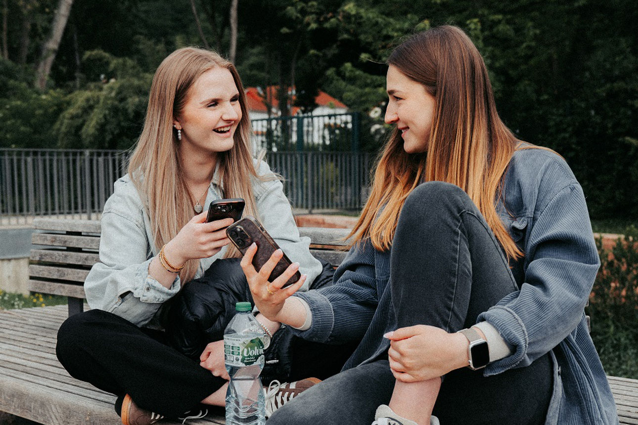 Zwei Personen sitzen auf einer Bank im Freien und schauen auf ihre Smartphones.