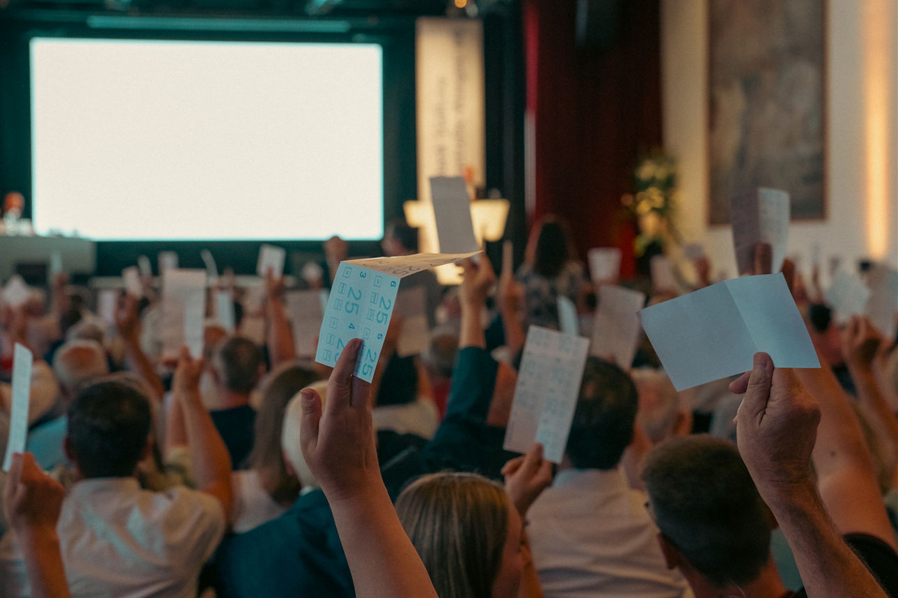 Publikum hebt Karten und Hände zur Abstimmung in einem Saal, vorne steht eine Bühne mit Leinwand