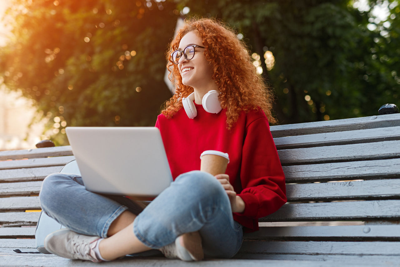 Mädchen sitzt mit Getränk auf einer Parkbank und hat einen offenen Laptop auf dem Schoß