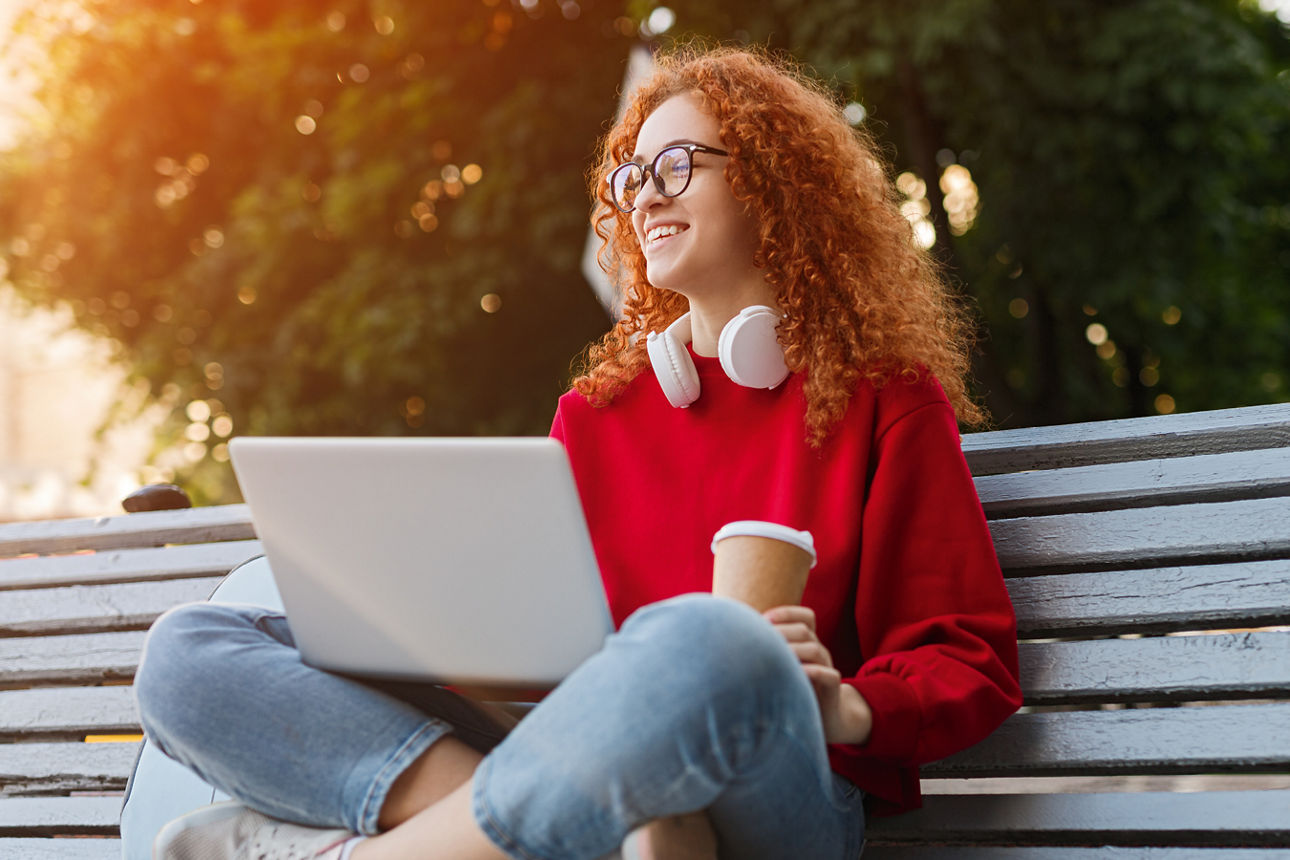 Mädchen sitzt mit Getränk auf einer Parkbank und hat einen offenen Laptop auf dem Schoß