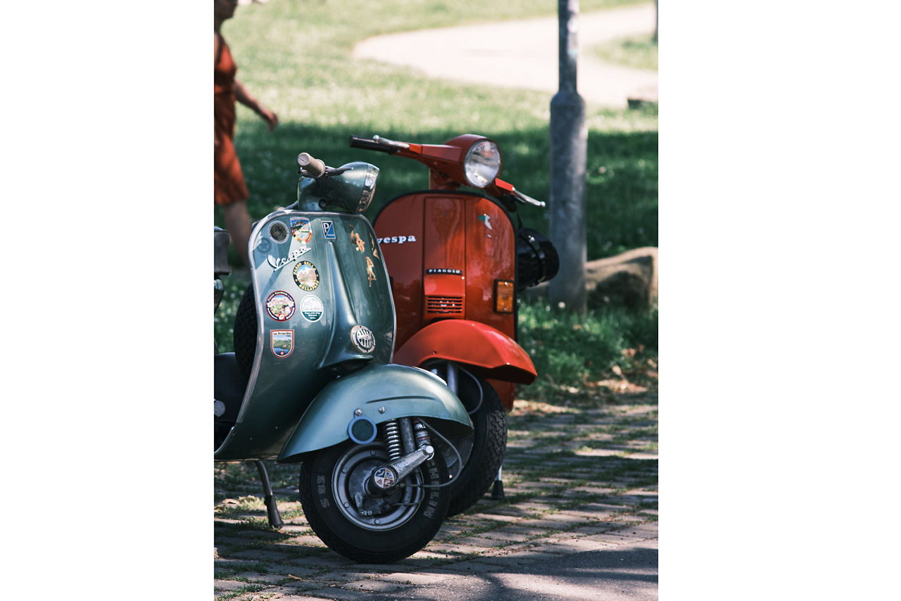 Zwei klassische Motorroller, einer hellblau mit mehreren Aufklebern, der andere rot, stehen auf einem gepflasterten Weg im Park.
