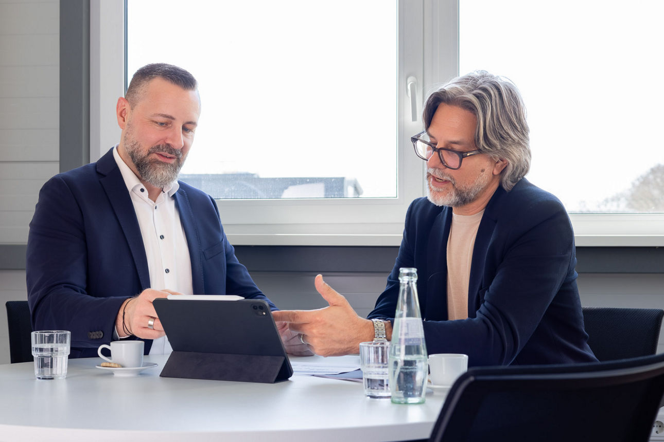 Zwei Personen besprechen Inhalte auf einem Tablet an einem Tisch; darauf stehen Wasserflaschen, Gläser und Tassen.