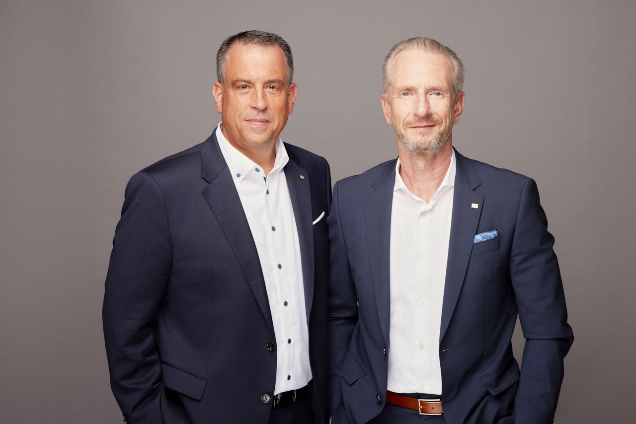 Zwei Männer in dunklen Anzügen stehen nebeneinander vor einem einfarbigen, grauen Hintergrund und blicken in die Kamera.