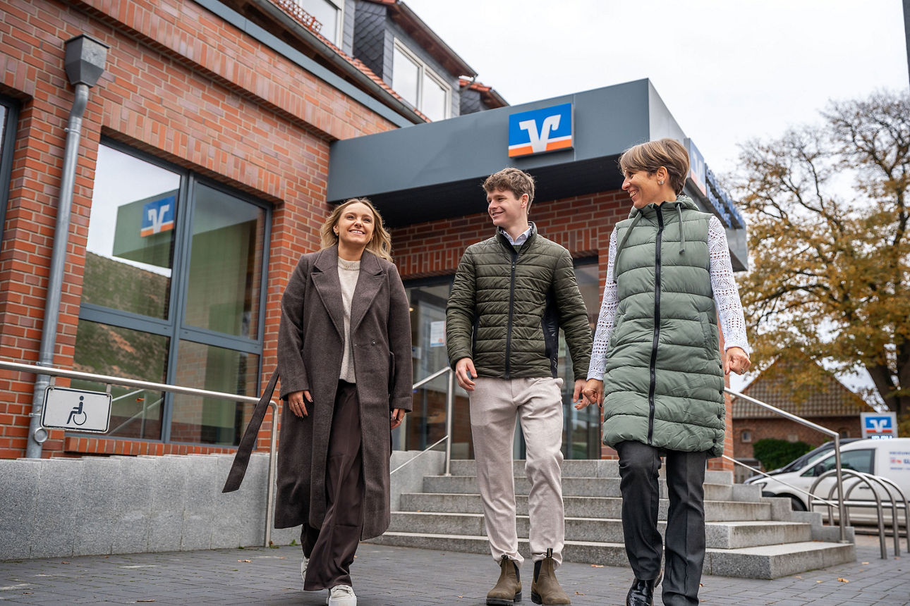 Drei Personen in winterlicher Kleidung gehen lächelnd an der Raiffeisen-Volksbank Neustadt eG vorbei