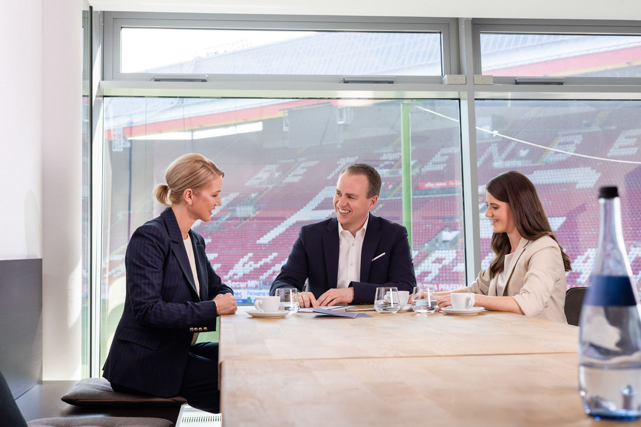 Drei Personen besprechen Unterlagen an einem Tisch; Gläser, Tassen und Dokumente liegen bereit, Stadion im Hintergrund.