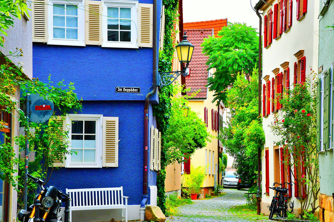 Blick in eine enge Gasse mit bunten Häusern, blaues Haus mit weißen Fensterläden und rotem Schriftzug, Kopfsteinpflaster, Fahrräder und grünem Bewuchs.