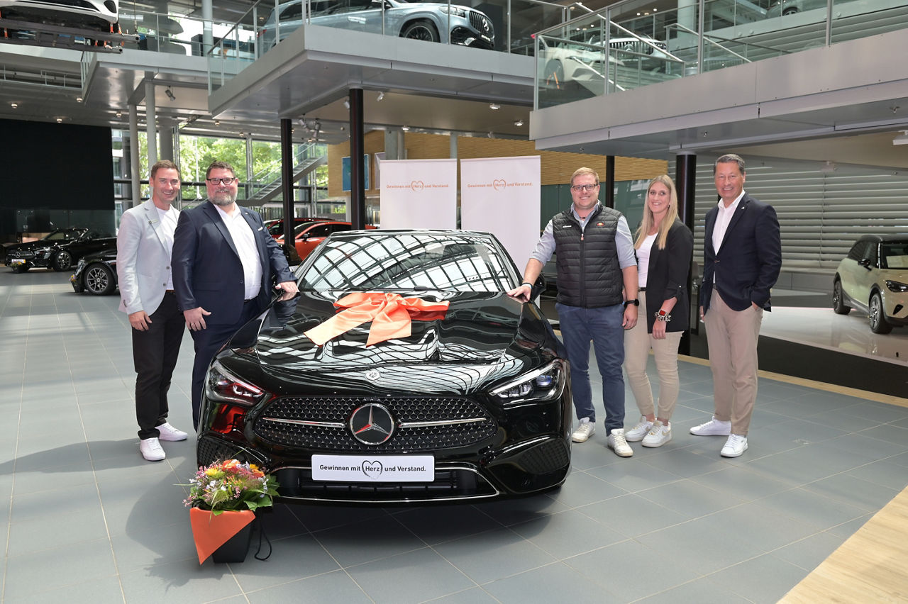Ein neuer Mercedes-Benz wird mit einer großen orangefarbenen Schleife und einem Blumenstrauß im Ausstellungsraum präsentiert.