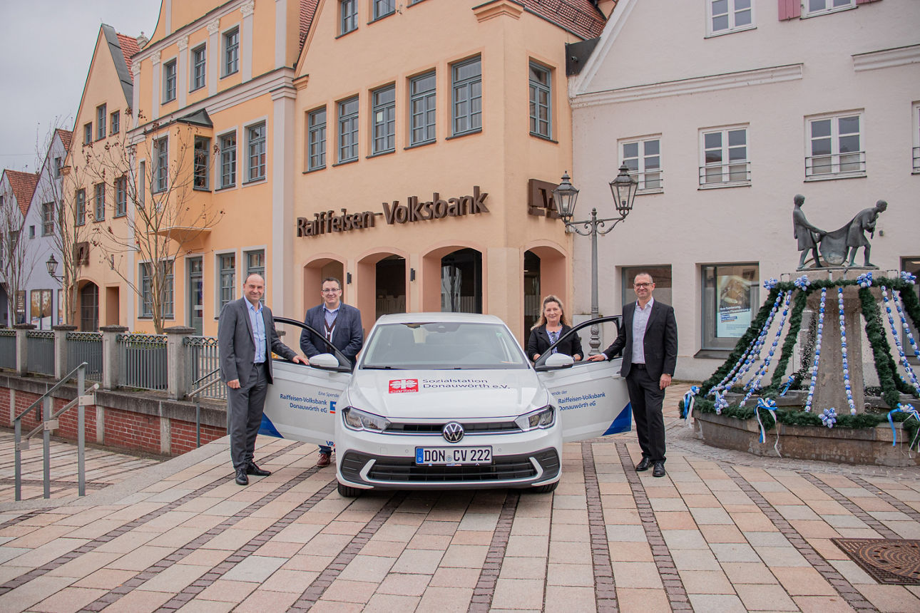 Vier Fachleute stehen neben einem Volkswagen mit Markenlogo vor einem Gebäude der Raiffeisen-Volksbank.