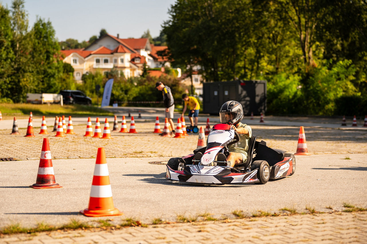 Kind fährt Gokart auf Outdoor-Strecke mit Verkehrskegeln, Zuschauer und Häuser im Hintergrund